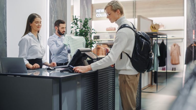 Clothing Store: Handsome Young Man At Checkout Counter Buys Clothes from Friendly Retail Sales Assistant, Paying with Contactless NFC Smartphone Touching Terminal